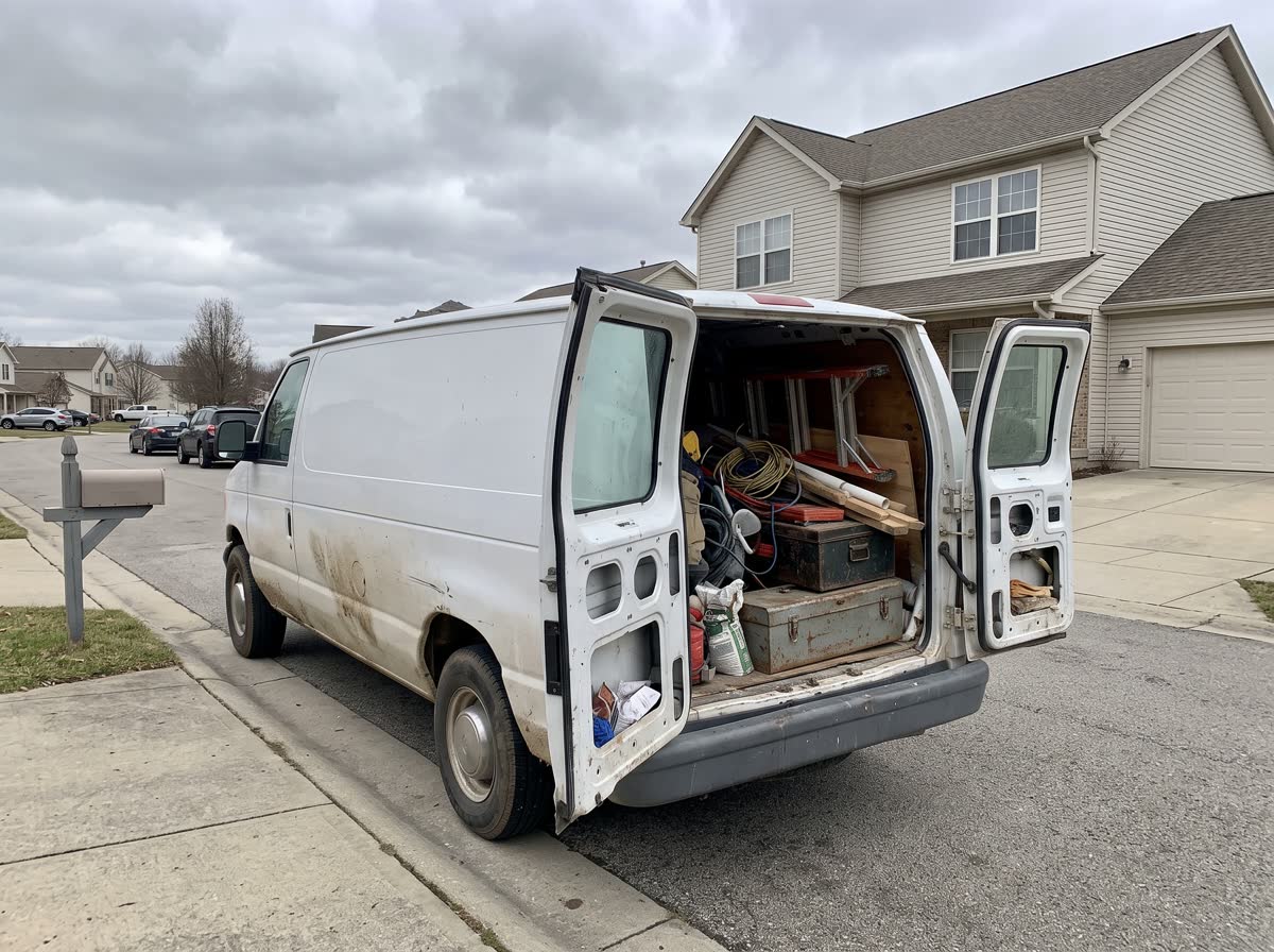 A working tradesman’s van parked at the kerb outside a suburban house, rear doors open on stacked tools and materials